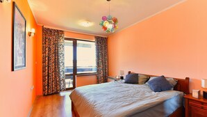 A bedroom with a large bed, a wooden nightstand, a ceiling light fixture, and a window with patterned curtains.