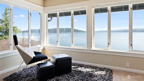 Una habitación con ventanas amplias que dan a un lago, una silla y dos otomanas sobre una alfombra estampada.
