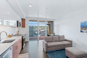 A modern kitchen with a sink, stove, and cabinets, a beige sofa, and a view of a balcony with a railing.