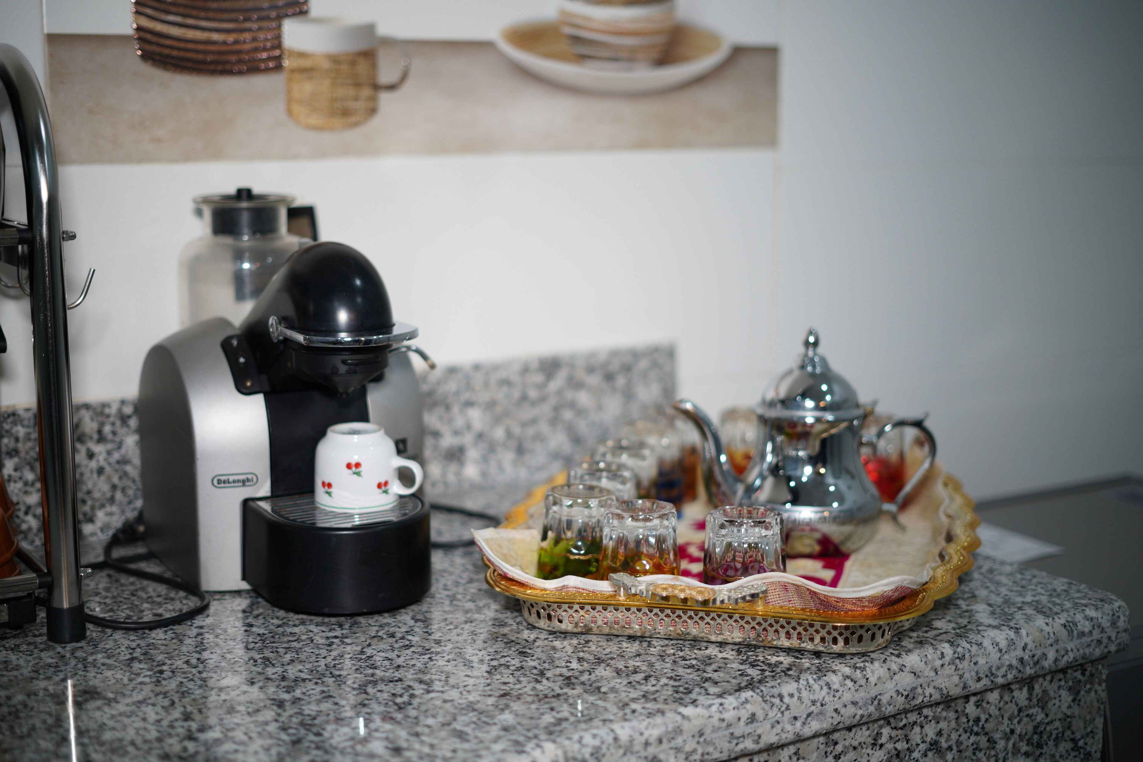 Uma cafeteira, um tabuleiro com copos e um bule sobre uma bancada de granito.