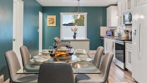 Una cocina moderna con armarios blancos, un refrigerador de acero inoxidable y un comedor con mesa de vidrio y sillas grises.