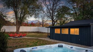 Un jacuzzi en un patio trasero con terraza de madera, valla blanca y árboles.