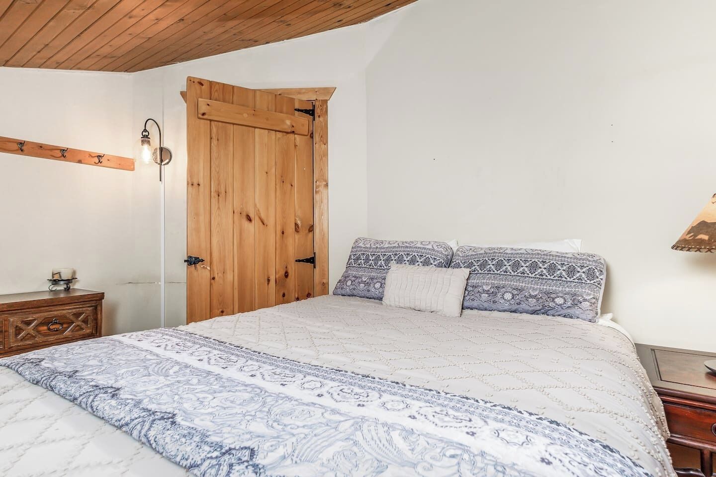 Un dormitorio con puerta de madera, cama con ropa de cama estampada, mesita de noche y lámpara de pared.