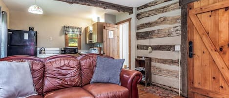 A cozy living room with a leather sofa, wooden walls, and a rustic kitchen in the background.