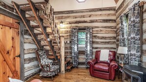 Interior de una cabaña de troncos con una escalera de madera, una chimenea, un sillón de cuero rojo y un sillón con estampado.