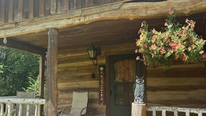Una cabaña de madera con un porche, un letrero de bienvenida, un banco y una maceta colgante de flores.
