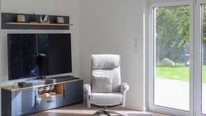 Un salon moderne avec un téléviseur à écran plat, un fauteuil gris et une porte-fenêtre coulissante donnant sur un jardin.