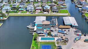 An aerial view of a marina with boats docked, a tennis court, and residential buildings in the background.