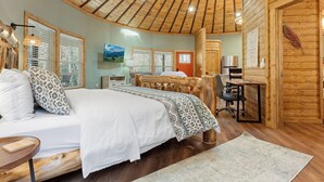 A cozy log cabin room with a bed, desk, and chair, wooden walls, and a ceiling with wooden beams.