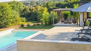 A swimming pool with a patio area, outdoor furniture, and a view of a forested hillside.