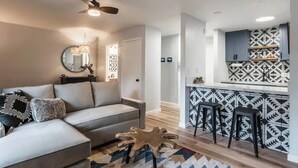 A modern living room with a grey sofa, a wooden coffee table, and a kitchen area with patterned tiles and bar stools.