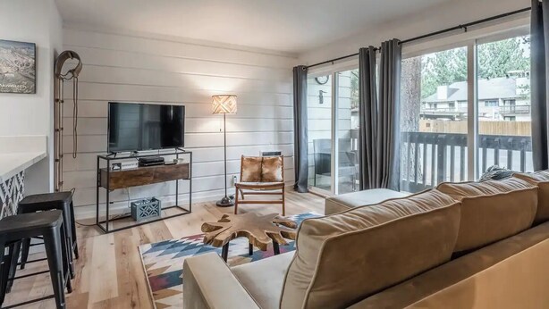 A modern living room with a flat-screen TV, a wooden coffee table, a patterned rug, and a large window with curtains.