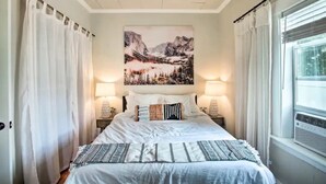 A bedroom with a bed, two bedside lamps, a mountain landscape painting, and a window with curtains. - Cheerful 3 bedroom Villa with Backyard Oasis (Spokane)