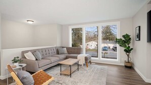 Ein Wohnzimmer mit Ecksofa, Couchtisch, gepolstertem Sessel, kleinen Beistelltisch und großem Fenster mit Blick auf die Straße.