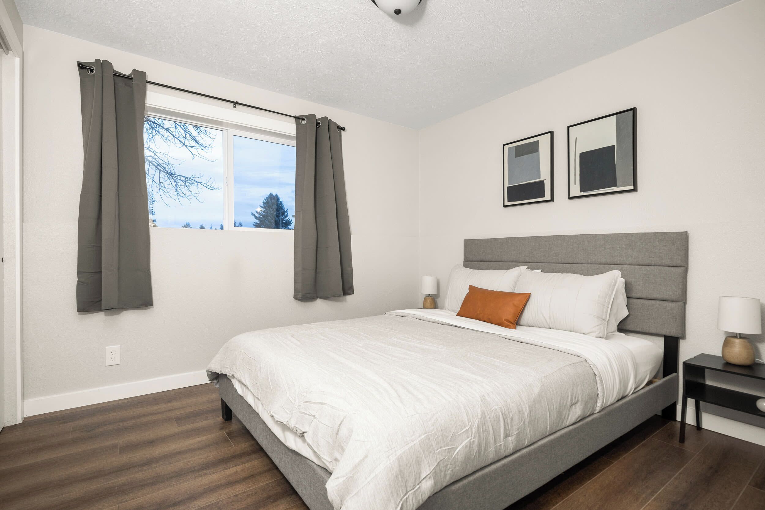 A bedroom with a bed, a nightstand, a window with curtains, and two framed pictures on the wall.