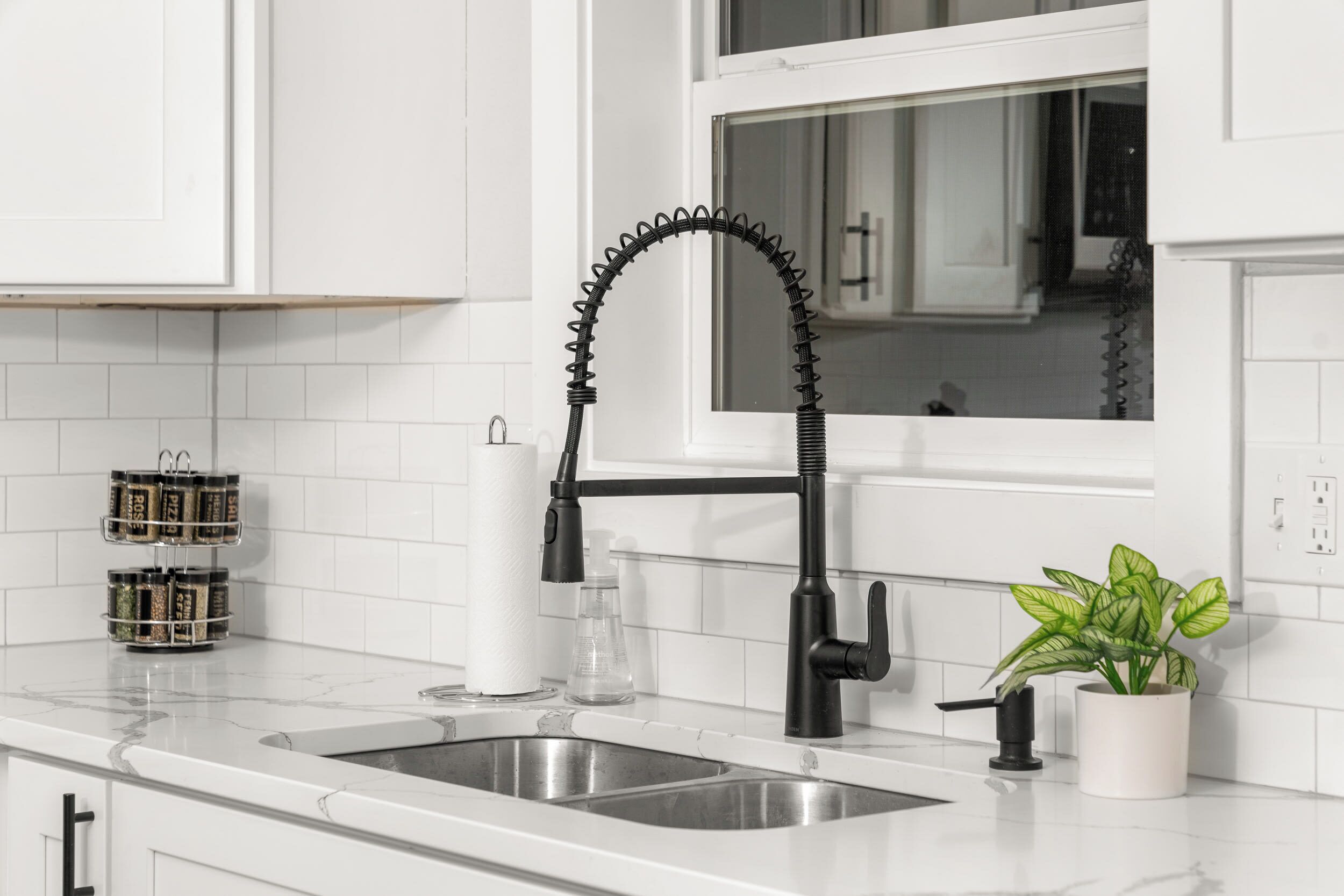 A kitchen with white cabinets, a black faucet, a sink, a window, and a potted plant.