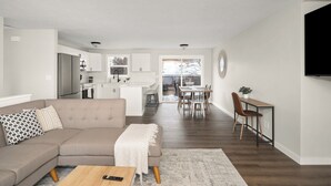 A modern living room with a beige sectional sofa, a wooden coffee table, and a dining area with a wooden table and chairs.