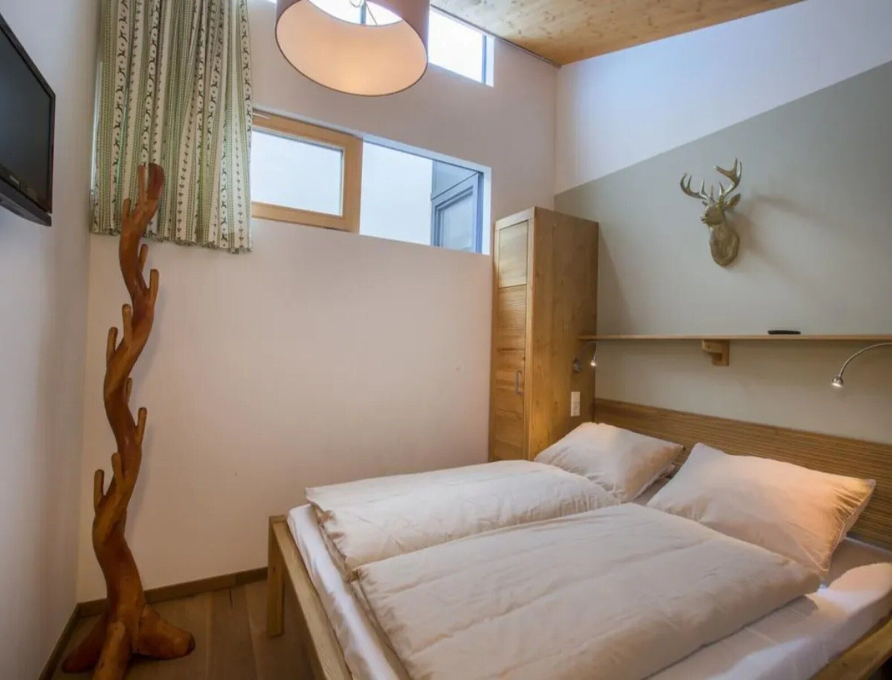 Ein Schlafzimmer mit einem Bett, einem hölzernen Kopfteil, einem aufgehängten Hirschkopf und einem Fenster mit Vorhängen.