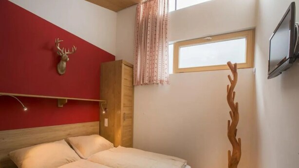 Un dormitorio con cama, armario de madera, cabeza de ciervo montada en la pared, ventana con cortinas y televisor en la pared.