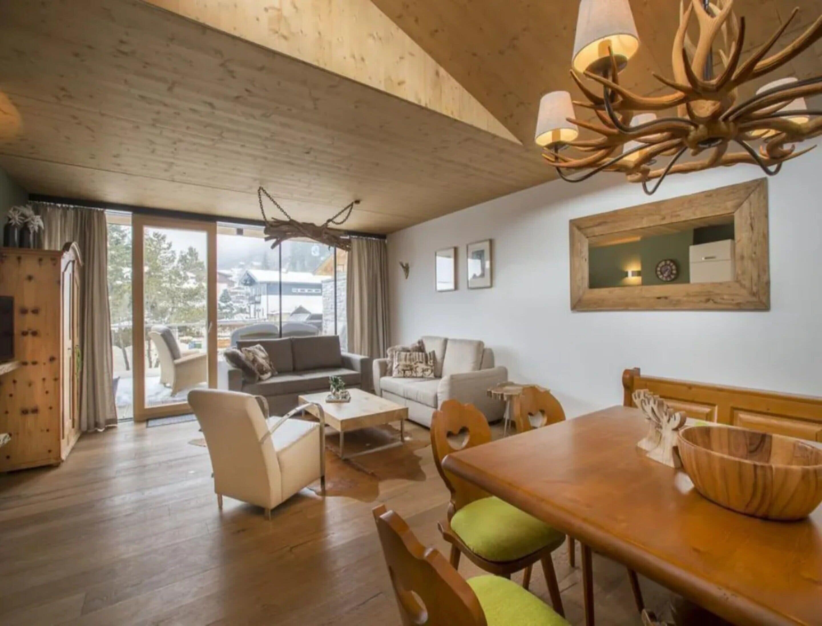 A modern living room with a wooden ceiling, a dining table with chairs, a sofa, and a large window with a view of a snowy landscape.