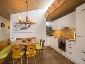 Un coin repas avec une table et des chaises en bois, un lustre, et une cuisine dotée de meubles blancs et d’appareils électroménagers.