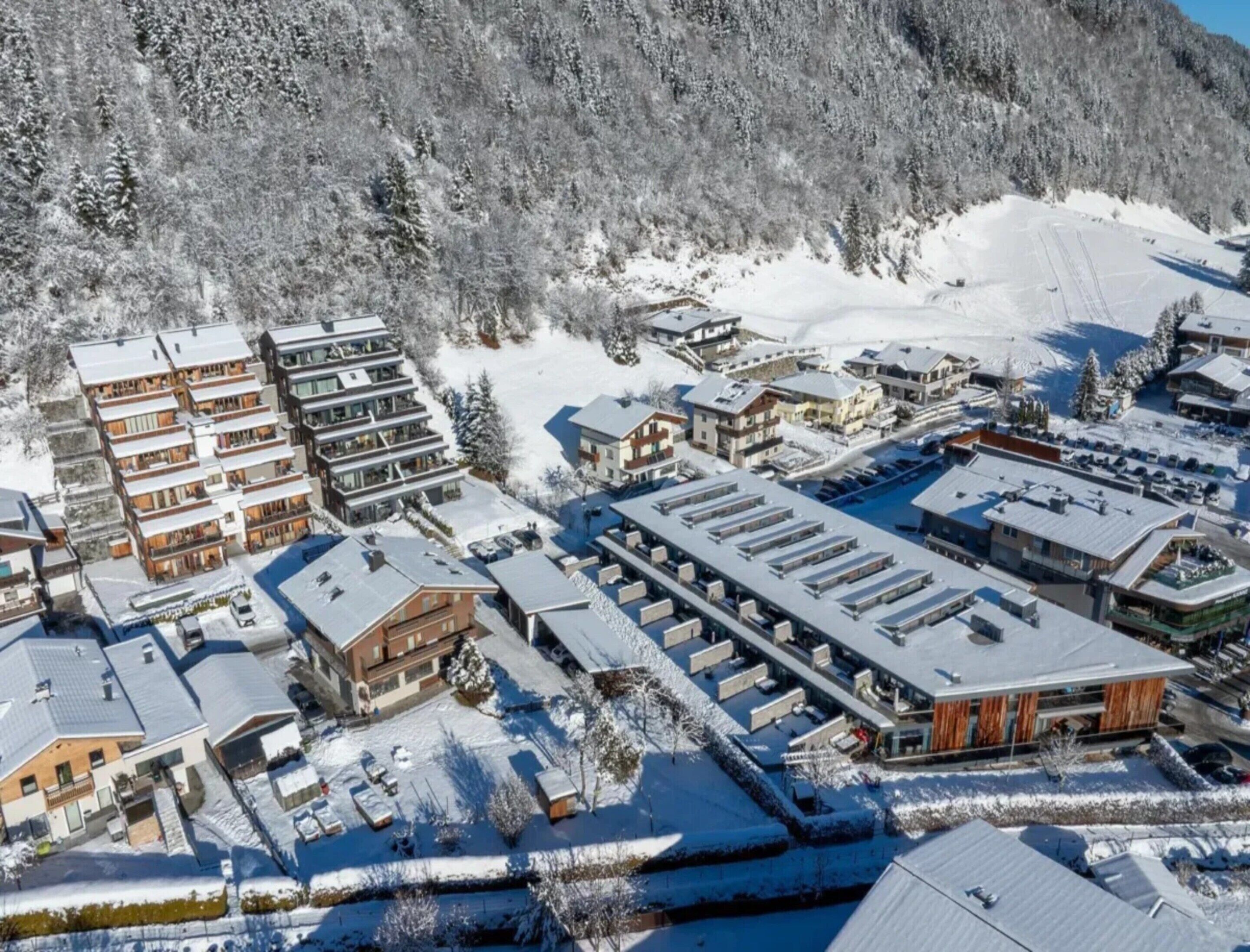 A snow-covered resort with multiple buildings, including a large hotel with a modern design and a ski lift visible in the background.