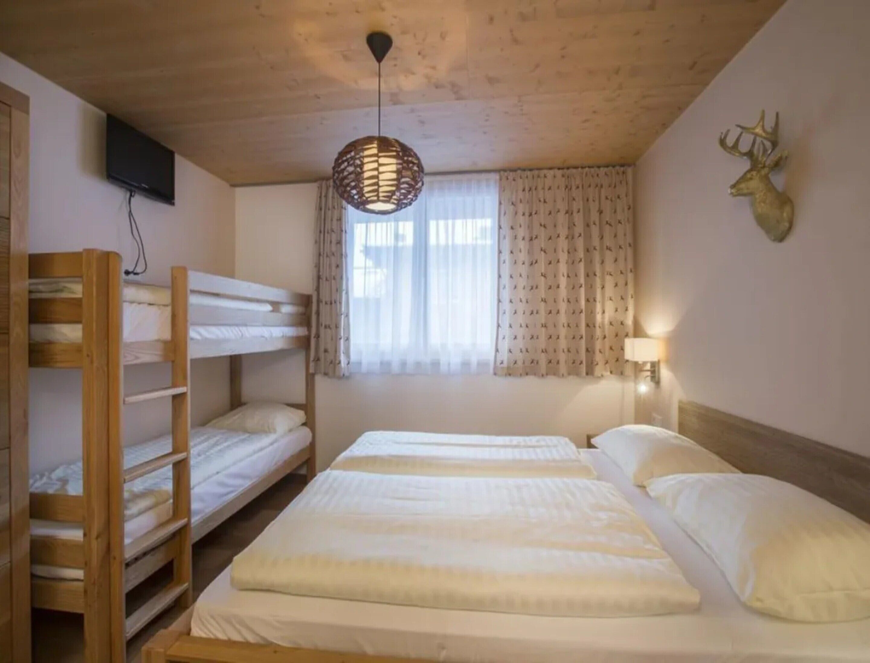 A room with bunk beds, a television mounted on the wall, a window with curtains, and a wall-mounted lamp.