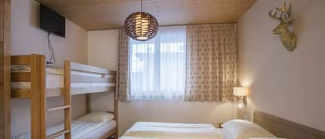 Une chambre avec des lits superposés, un téléviseur fixé au mur, une fenêtre avec des rideaux et une lampe fixée au mur.