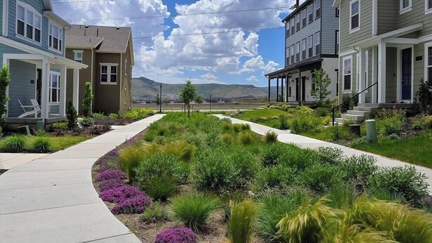 A residential street with two-story houses, a landscaped median, and a clear blue sky. - Skip Rock Retreat | Near SLC | Cozy Mid-Term Stay (South Jordan)