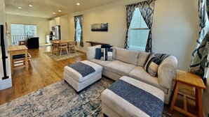A spacious living area with a large sectional sofa, ottomans, a dining table with chairs, and a kitchen in the background. - Skip Rock Retreat | Near SLC | Cozy Mid-Term Stay (South Jordan)