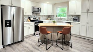 Uma cozinha moderna com armários brancos, eletrodomésticos de aço inoxidável, uma ilha central com dois banquetes e uma janela com vista para vegetação.