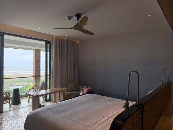 A modern hotel room with a large bed, a ceiling fan, a view of the ocean, and a balcony with outdoor furniture.