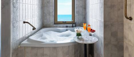 A modern bathroom with a large bathtub, a window with a view of the sea, and a small table with drinks and flowers.