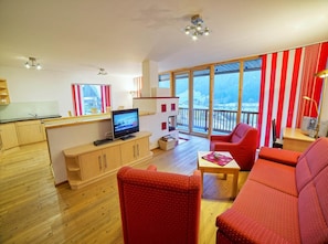 A modern living room with a television, red sofas, wooden flooring, and large windows offering a view of a snowy landscape. - Zirbenholz Alpine Maisonette Stay (Wald im Pinzgau)