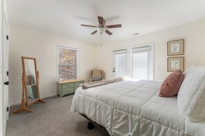 Une chambre à coucher comprenant un lit, un ventilateur de plafond, une fenêtre avec des stores, une commode et un miroir.