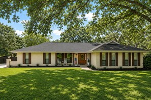 Une maison de plain-pied avec un porche à l’avant, un petit jardin devant et un grand arbre en arrière-plan.