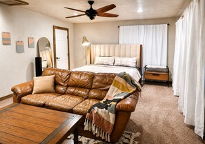 A bedroom with a bed, a leather sofa, a wooden coffee table, a ceiling fan, and a window with curtains.