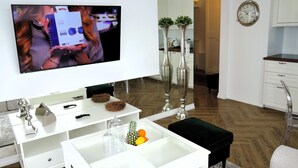 A modern living room with a flat-screen TV, white furniture, a glass coffee table with fruits, and a kitchen area in the background. - Comfortable Apartment With a sea View (Miedzyzdroje)