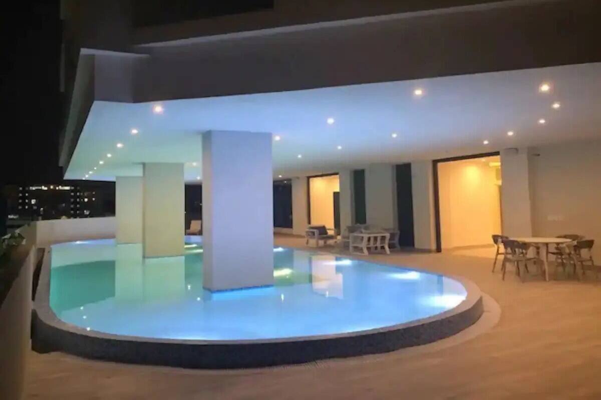 A modern outdoor swimming pool area with a circular design, surrounded by a wooden deck. There are several seating areas with tables and chairs, and the space is illuminated by recessed ceiling lights.
