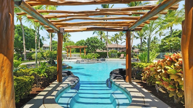 Zona piscina con una pergola in legno, circondata da piante tropicali e palme.