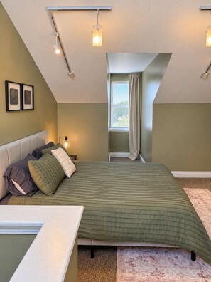 A bedroom with a sloped ceiling, a bed with a green bedspread, a nightstand with a lamp, and a window with curtains.