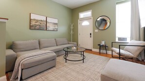 A living room with a sofa, ottoman, coffee table, and a chair. There is a door, a coat rack, and a window with curtains. - Spacious 4-bedroom townhome in St. Louis  (St. Louis)