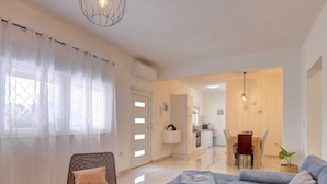 Ein modernes Wohnzimmer mit einem grauen Sofa, einem Holzstuhl und einem weißen Couchtisch. Im Hintergrund ist ein Essbereich mit Tisch und Stühlen zu sehen.