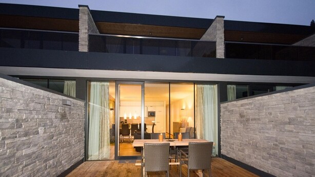 Ein modernes Haus mit Holzterrasse, Steinwänden und einer Glastür, die zu einem Essbereich führt.