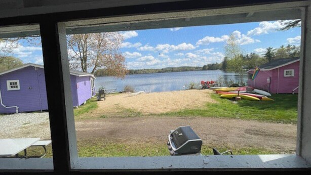 Vista a través de una ventana de una propiedad frente al lago, con una zona de arena, embarcaciones y dos edificios morados.