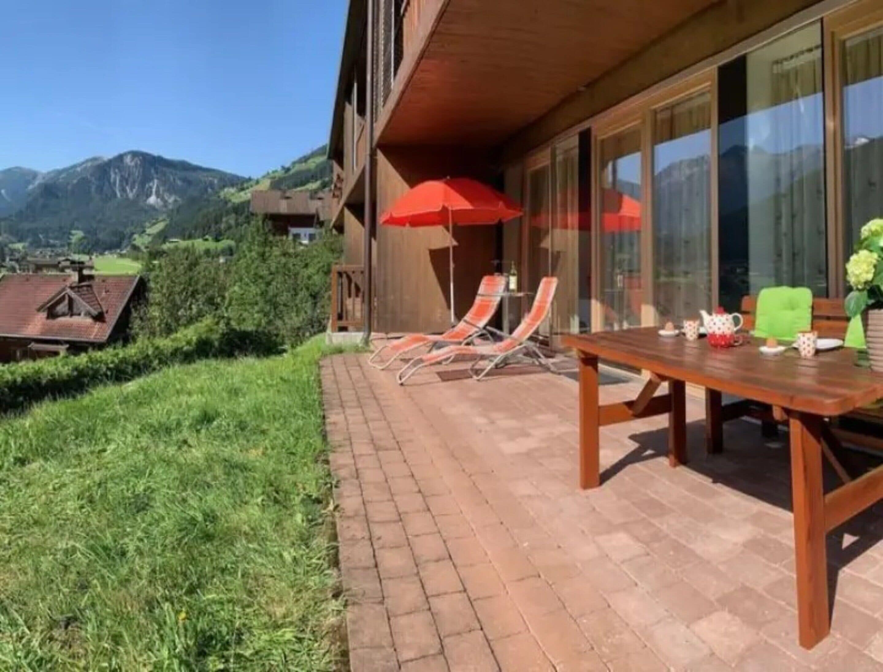 A patio with a table and chairs, a red umbrella, and a view of mountains.