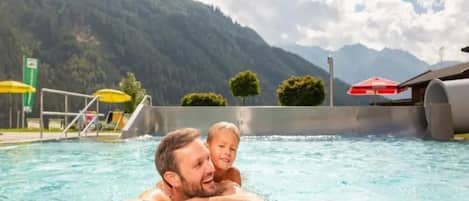 A swimming pool with a mountain backdrop, a diving board, and umbrellas.
