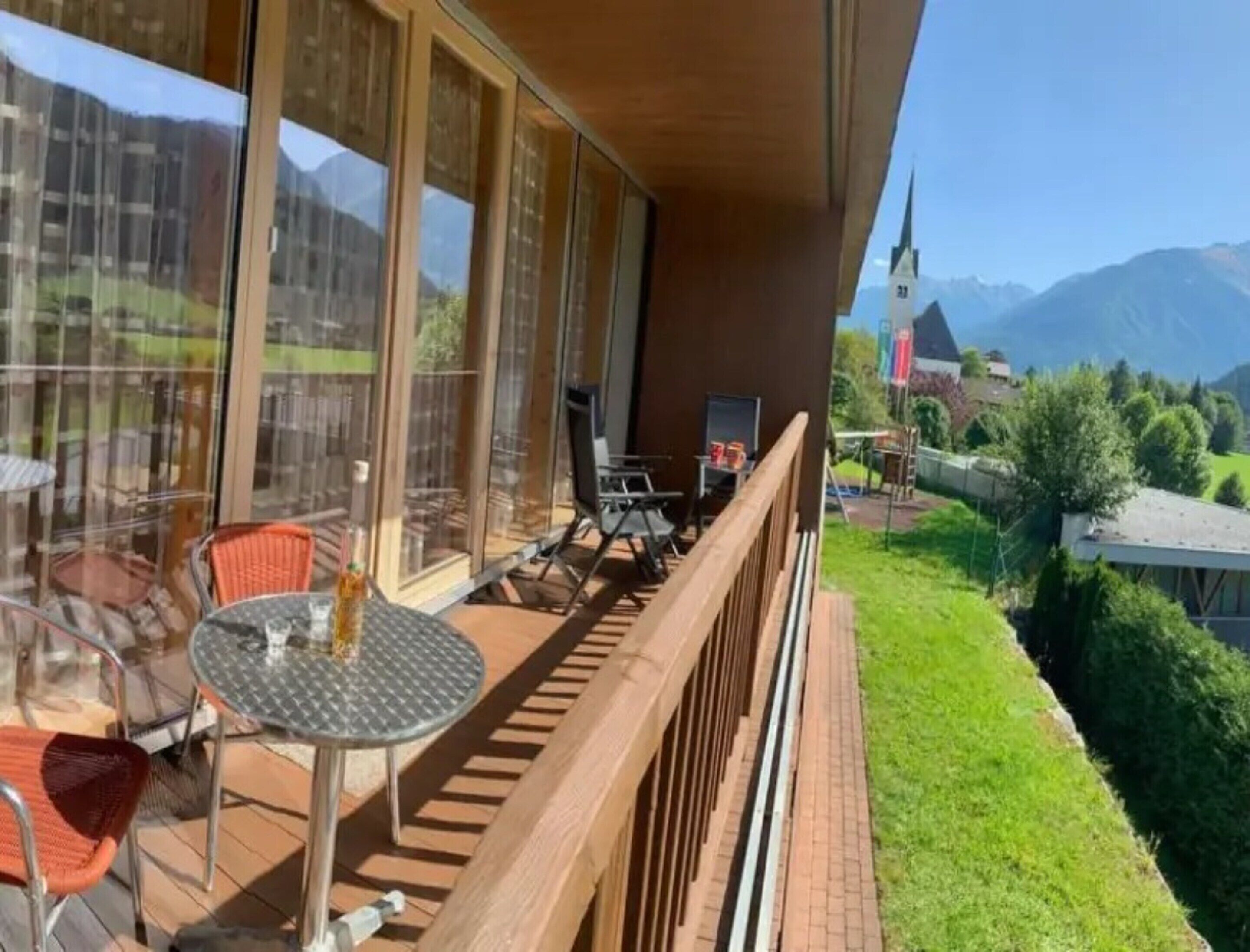 A balcony with a table and chairs, overlooking a mountainous landscape with a church.