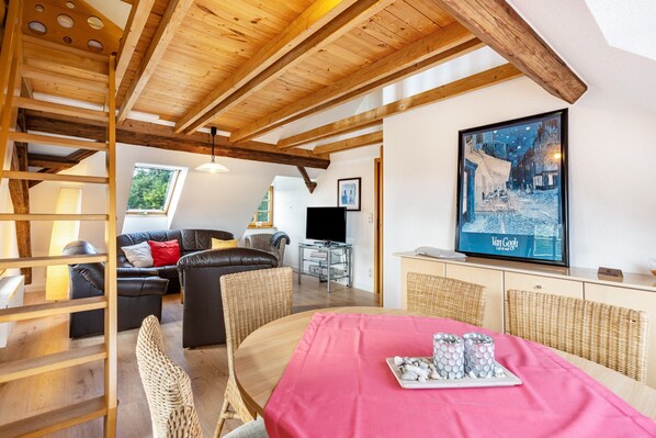 A cozy living room with a wooden ceiling, a dining area with a pink tablecloth, and a television displaying a winter scene.