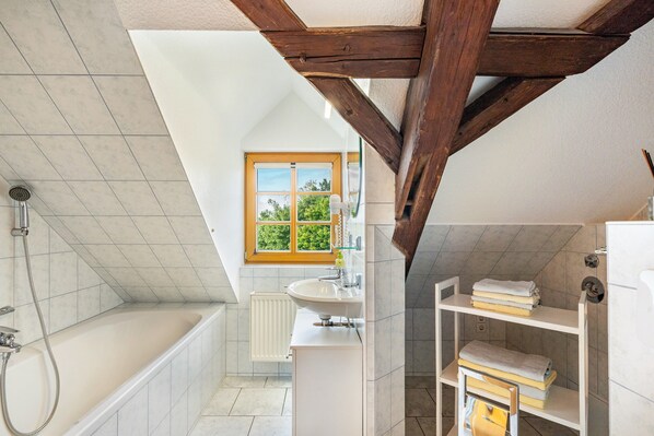 Un baño moderno con bañera, ducha y lavamanos, vigas de madera, una ventana con vistas y un estante con toallas.